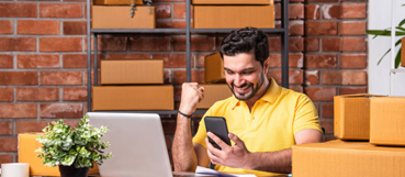 Smiling small business owner in a shipping workspace checking a smartphone while working at a laptop surrounded by shipping boxes, celebrating a successful online sale.