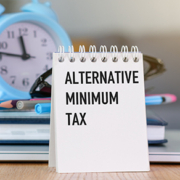 Desk notepad reading "Alternative Minimum Tax" in front of a blue alarm clock, representing year-end tax planning with a CPA in Columbia, MD at Weyrich, Cronin & Sorra.