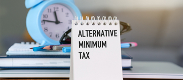 Desk notepad reading "Alternative Minimum Tax" in front of a blue alarm clock, representing year-end tax planning with a CPA in Columbia, MD at Weyrich, Cronin & Sorra.